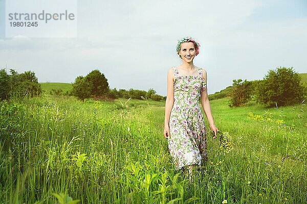 Junges glückliches lächelndes Mädchen in einem Kattunkleid mit einem Blumenstrauß geht eine Landstraße mit grünem Gras entlang