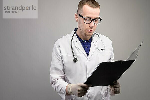 Porträt eines jungen männlichen Arztes mit Brille und einem Stethoskop um den Hals. In den Händen ein Ordner  ein Tablet Halter für Papiere. Studioaufnahme auf grauem Hintergrund