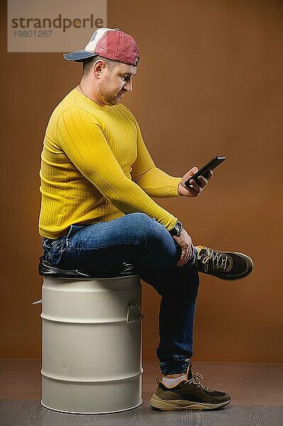 Ein molliger Mann in Freizeitkleidung und mit Mütze sitzt auf einem Metallfass und hält ein Telefon in der Hand. Schreibt SMS oder prüft seine Post. Studioporträt