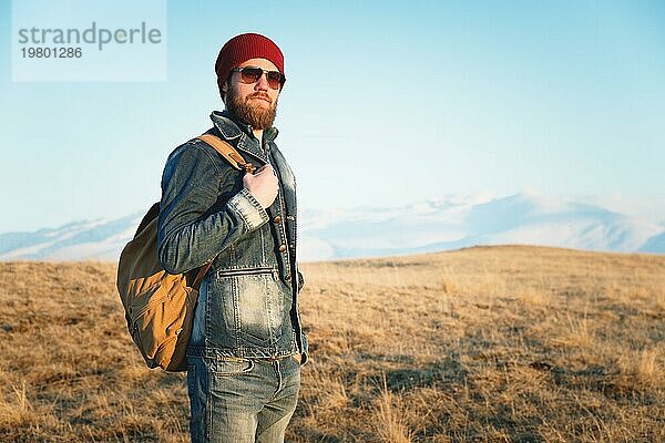 Porträt eines nachdenklichen stilvollen Hipsters mit Bart  Sonnenbrille und Hut mit einem Rucksack vor dem Hintergrund des schneebedeckten Berges Elbrus im Kaukasus bei Sonnenuntergang