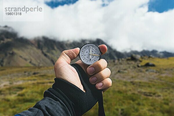 Blickpunktaufnahme. Eine Ichperspektive eines Mannes  der einen Kompass vor dem Hintergrund einer epischen Landschaft mit Klippen  Hügeln und einem blaün Himmel mit Wolken hält