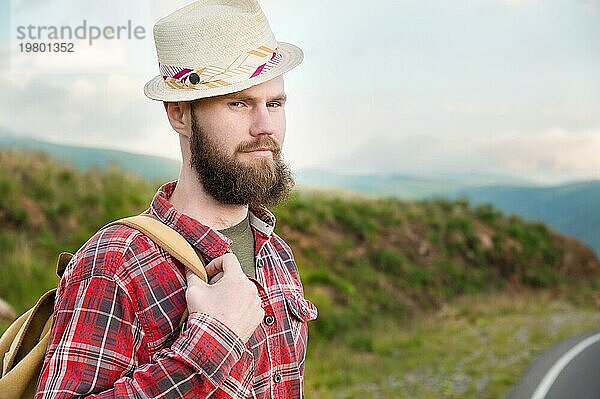 Attraktiver  romantischer  bärtiger  kaukasischer Mann mit Hut  kariertem Hemd und einem Rucksack. Steht auf dem Lande in den Bergen und schaut in die Kamera. Raum kopieren. Per Anhalter