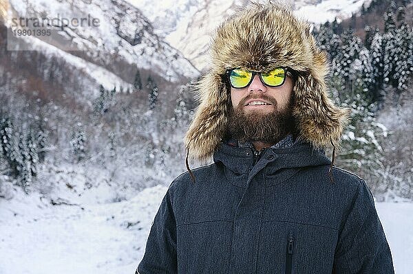 Porträt eines attraktiven jungen bärtigen Mannes mit einer großen Pelzmütze und einer Sonnenbrille vor einem verschneiten Wald in den Bergen