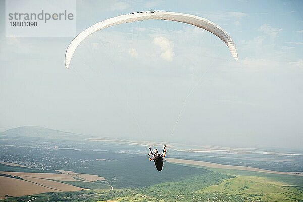 Professioneller Gleitschirmflieger in einem Kokon Anzug fliegt hoch über dem Boden gegen den Himmel und die Felder