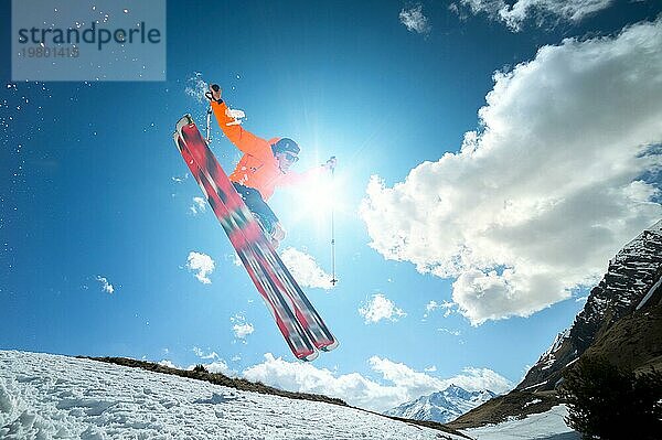 Ein junger stilvoller Mann mit Sonnenbrille und Mütze führt einen Trick beim Springen mit einem Kicker aus Schnee gegen den blaün Himmel und die Sonne an einem sonnigen Tag aus. Das Konzept der Park Skifahren im Winter oder Frühling