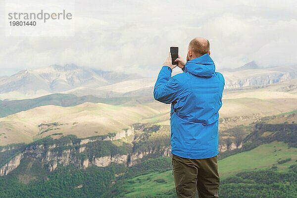 Ein bärtiger Mann in einer membranblauen Jacke steht in der Natur vor der Kulisse der Berge und fotografiert die Telefonlandschaft