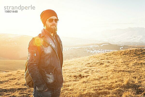 Porträt eines nachdenklichen stilvollen Hipsters mit Bart  Sonnenbrille und Hut mit einem Rucksack vor dem Hintergrund des schneebedeckten Berges Elbrus im Kaukasus bei Sonnenuntergang