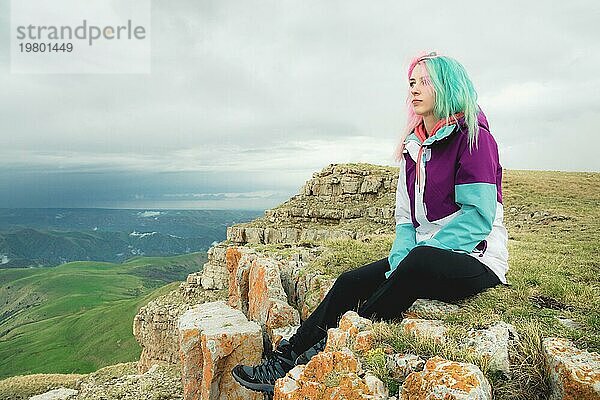 Eine Reisende mit buntem Haar sitzt am Rande einer Klippe und blickt auf den Horizont vor dem Hintergrund einer felsigen Hochebene