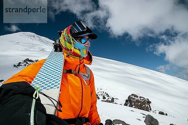 Weitwinkelaufnahme eines jungen  großgewachsenen Profi Skifahrers  der seine Skier hoch in den Bergen hält und auf die schneebedeckten Gipfel blickt. Sportler mit einem Rucksack und in Schutzkleidung in den Bergen beim Freeriden