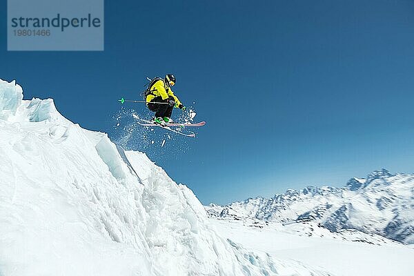 Ein Skifahrer in voller Sportausrüstung springt von der Spitze des Gletschers in den Abgrund vor dem Hintergrund des blaün Himmels und der schneebedeckten Berge des Kaukasus. Elbrus Gebiet. Russland