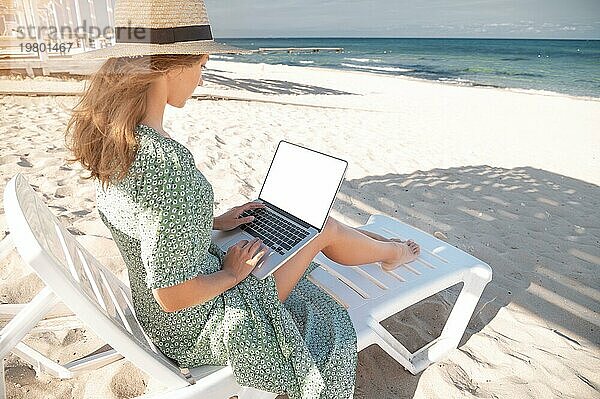 Rückansicht einer attraktiven jungen kaukasischen Frau in grünem Kleid und Hut  die im Schatten eines hölzernen Sonnenschirms auf einer Liege mit einem Laptop in den Händen sitzt. Ausgeschnittener Laptopbildschirm