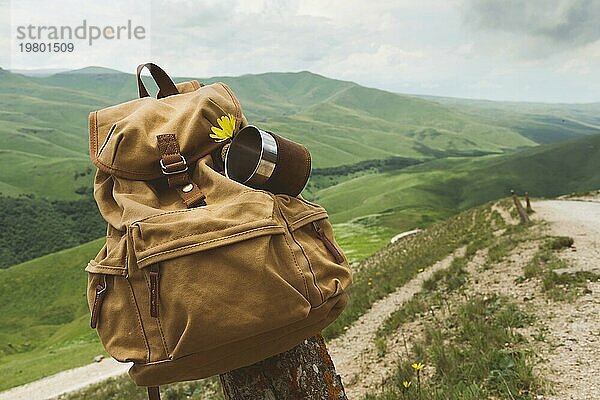 Hipster gelben Vintage Rucksack mit einem Becher auf sie mit einem Becher Nahaufnahme Vorderansicht befestigt. Reisetasche des Reisenden in den Hintergrund einer Berglandschaft und einer Straße