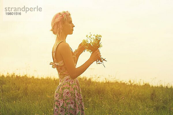 Junge Frau im Kleid mit Blumenstrauß in den Händen bei Sonnenuntergang auf dem Feld. Getöntes warmes Bild