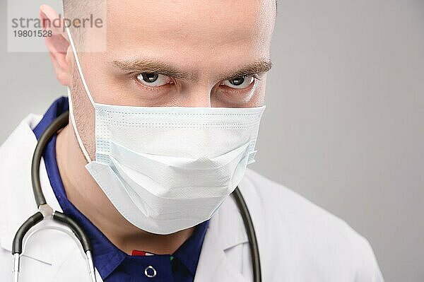 Ein junger  weißer  wütender Arzt in Maske und Kittel blickt bedrohlich in die Kamera. Verschwörung durch Impfung