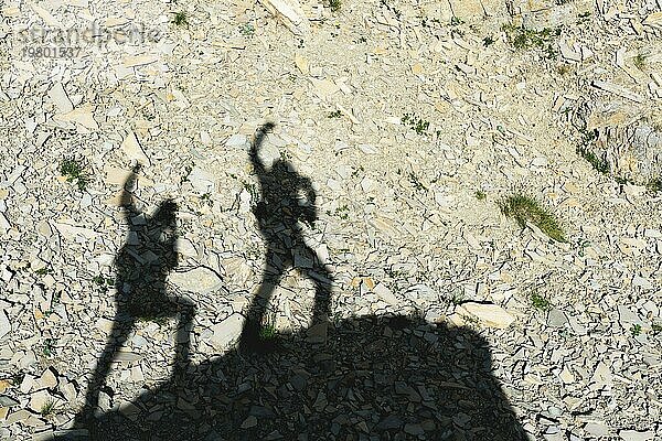 Zwei Schatten von Fotografen auf der Erde  die mit erhobenen Händen bergauf gehen und über Sieg und Erfolg schreien. Silhouetten von zwei Menschen  die bergauf gehen
