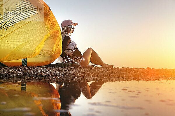Attraktive stilvolle junge kaukasische Frau in Kappe Sonnenbrille und Kitesurfer Outfit sitzt auf dem sandigen Ufer neben ihrem Drachen bei Sonnenuntergang spiegelt sich im Wasser