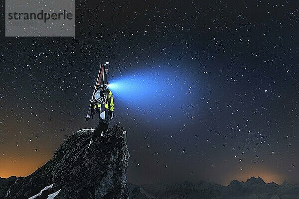 Nächtliche Landschaft. Ein professioneller Skitourengeher mit Rucksack und Skiern steht auf einem Felsen in den Bergen und leuchtet mit seiner Stirnlampe in den Himmel
