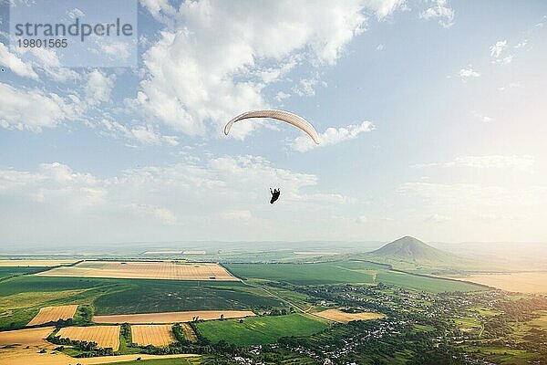 Professioneller Gleitschirmflieger in einem Kokon Anzug fliegt hoch über dem Boden gegen den Himmel und Felder mit Bergen