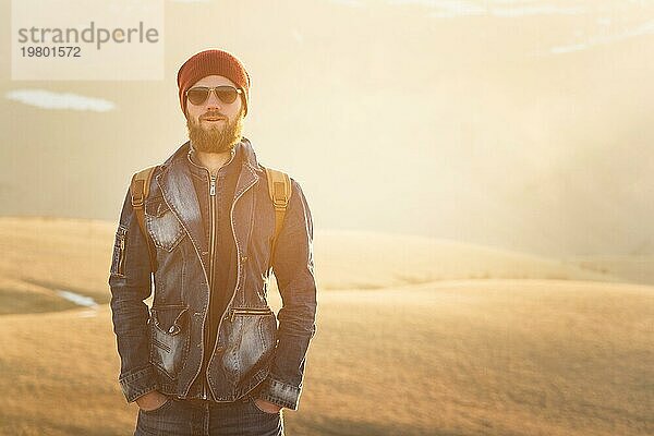 Modeporträt eines lächelnden bärtigen Hipster jungen Mannes mit Sonnenbrille  einen Rucksack und Hut auf einem Hintergrund mit copyspase in den Bergen bei Sonnenuntergang  Ein selbstbewusster Mann mit einem Bart