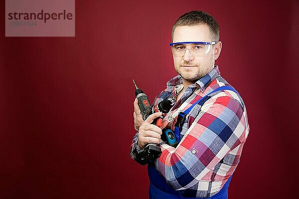 Ernster kaukasischer Mechaniker mit Schutzbrille  der Holzbearbeitungswerkzeuge hält. Schreinerporträt auf rotem Studiohintergrund. Kopierraum