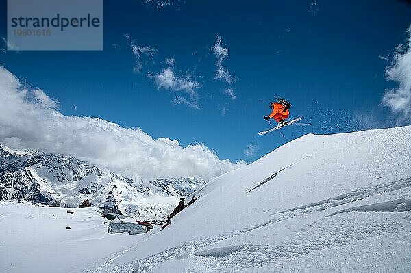 Ein junger männlicher Profi Skifahrer in einem orangefarbenen Skianzug fliegt über die Berge  nachdem er von schneebedeckten Felsvorsprüngen gesprungen ist. Freeride Sportgemeinschaft vor der Kulisse schneebedeckter Gipfel