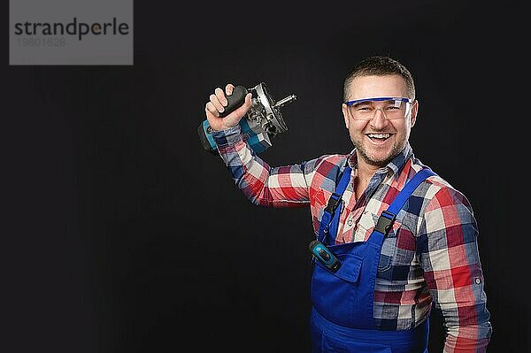 Porträt eines attraktiven Mechanikers in Uniform und kariertem Hemd mit Bartstoppeln im Gesicht  der ein Gerät in der Hand vorführt  Studioaufnahme  er lacht und hält einen Elektroschneider in der Hand