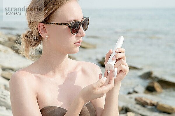 Eine junge kaukasische Frau drückt an einem sonnigen Tag am Strand Sonnencreme aus einer Tube. UV Schutz