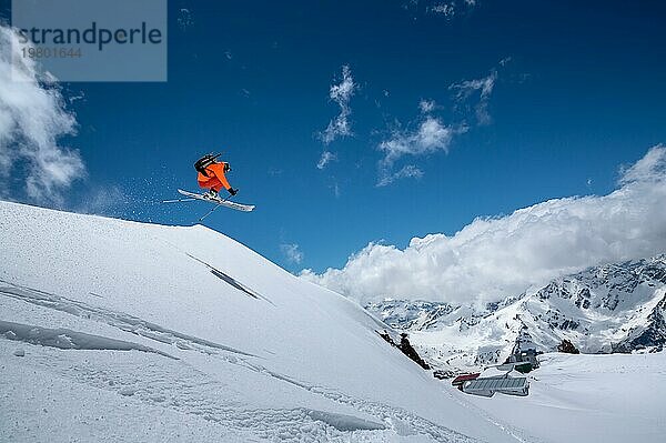 Fliegender Sprung über eine Piste Freestyle Skifahrer im orangefarbenen Skianzug in den verschneiten Bergen an einem sonnigen Tag