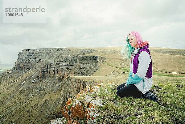 Eine Reisende mit buntem Haar sitzt am Rande einer Klippe und blickt auf den Horizont vor dem Hintergrund einer felsigen Hochebene