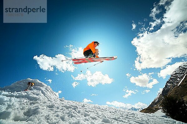 Ein junger stilvoller Mann mit Sonnenbrille und Mütze führt einen Trick beim Springen mit einem Kicker aus Schnee gegen den blaün Himmel und die Sonne an einem sonnigen Tag aus. Das Konzept der Park Skifahren im Winter oder Frühling