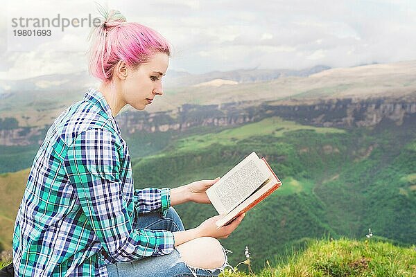 Ein reisendes Mädchen sitzt in den Bergen im Gras und liest ein Buch vor dem Hintergrund epischer Berge. Das Konzept des Lesens während der Erholung und des Urlaubs