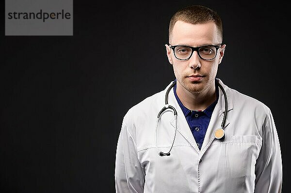 Ein Studioporträt eines attraktiven jungen kaukasischen Arztes in weißem Kittel und mit Brille und einem Stethoskop um den Hals. Steht und schaut in die Kamera. Auf schwarzem Hintergrund