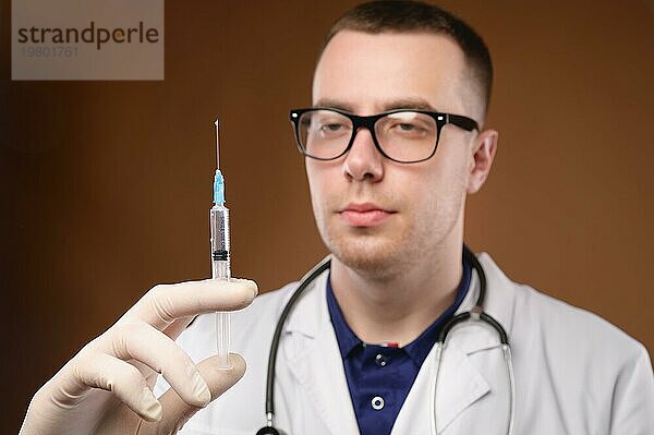 Junger kaukasischer Arzt  der sich darauf vorbereitet  eine Impfung mit einer Spritze durchzuführen. Studioaufnahme auf einem braunen Hintergrund. Lassen Sie Luft aus der Spritze vor der Injektion
