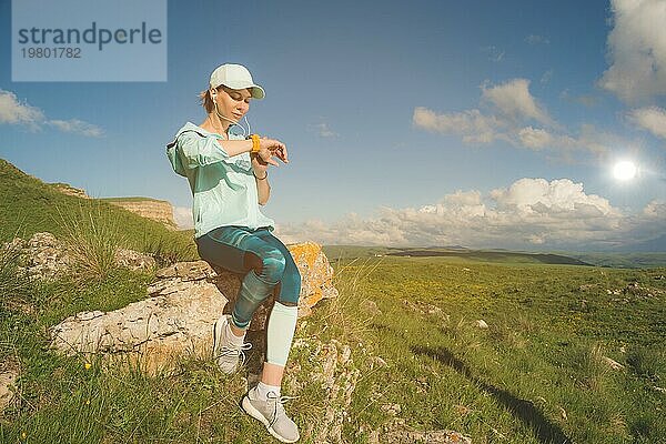 Porträt eines lächelnden jungen Fitness Mädchens mit Mütze und Kopfhörern  das auf einem Felsen sitzend im Freien vor einem blaün Himmel mit Wolken ihre Smartwatch überprüft