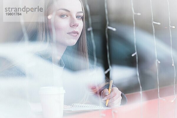 Porträt einer attraktiven jungen Journalistin mit Stift und Notizblock in einem Café hinter einer Vitrine. Kontrastlose Ansicht durch die Spiegelung des Fensterglases mit Blick in die Kamera