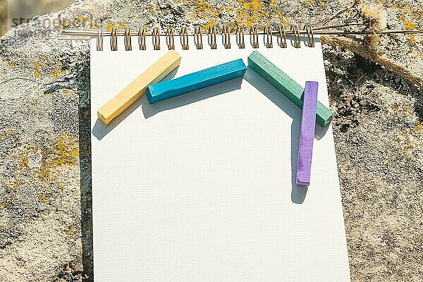 Nahaufnahme Kunstwerk für den Designer. Ein leerer Notizblock für die Zeichnung Pastell  auf dem eine künstlerische Pastellkreiden Farbe Kreiden von lila grün blau und gelb liegt auf einem Felsen in der Natur im Freien. Das Konzept der Kreativität und der Einheit mit der Natur