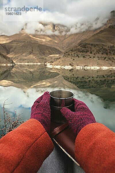 Eine Ichperspektive von Frauenhänden in Mänteln und Fäustlingen  die einen Metallbecher mit Tee oder Kaffee und ein ledernes Taschenkalenderbuch vor dem Hintergrund eines Bergsees und Felsen in den Wolken halten. Reisekonzept