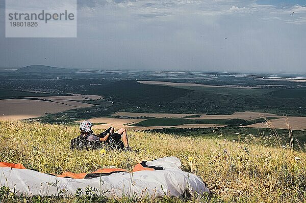 Ein professioneller Gleitschirmflieger in voller Ausrüstung und mit Helm liegt auf dem Gras hoch in den Bergen und schaut in die Wolken