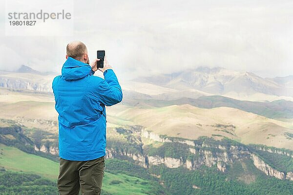 Ein bärtiger Mann in einer membranblauen Jacke steht in der Natur vor der Kulisse der Berge und fotografiert die Telefonlandschaft