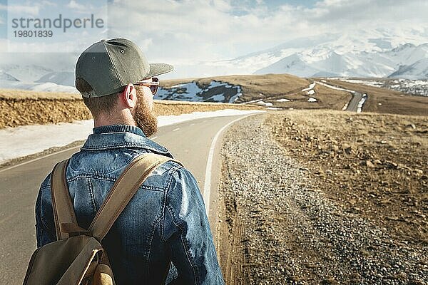 Ein bärtiger Mann mit einer Mütze und einem Rucksack  der bereit ist  einen langen Weg zu gehen. Ein Mann auf einer Landstraße vor der Kulisse von Bergen und Wolken