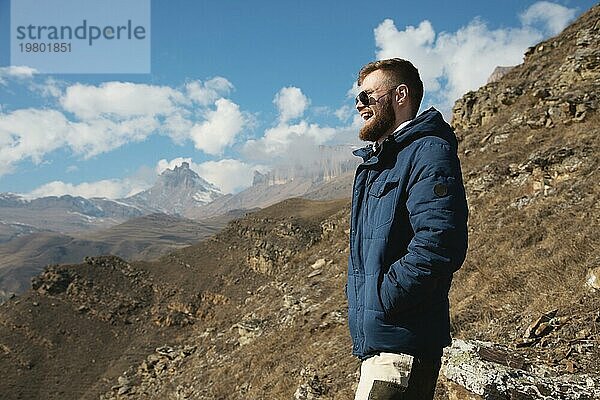 Hipster Reisender in Daunenjacke und Sonnenbrille steht an einem Berghang vor der Kulisse epischer Felsen und lächelt. Das Konzept  das Glück in den Bergen zu finden