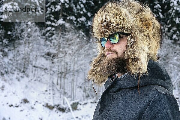 Porträt eines attraktiven jungen bärtigen Mannes mit einer großen Pelzmütze und einer Sonnenbrille vor einem verschneiten Wald in den Bergen