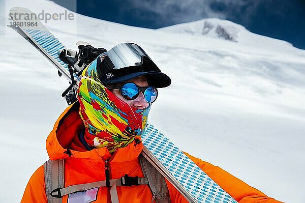 Bergporträt eines professionellen Freeride Skifahrers in orangefarbener Kleidung mit Skistöcken und Skiern auf den Schultern. Steht hoch in den Bergen auf einer verschneiten Piste