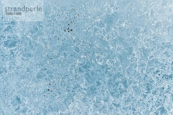 Nahaufnahme der Oberfläche eines Gletscherfragments mit einer Struktur aus Streifen und Blasen  die mit Sand und felsigem Gestein durchsetzt ist. Eisblaue Textur im Licht. Kleine Tiefenschärfe