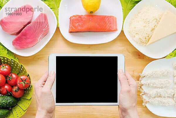 Design Werkstück. Weibliche Hände halten einen Tablet PC über einen Tisch mit rohen Meeresfrüchten  Käse und Gemüse. Blank Display für die Präsentation zum Thema Lebensmittel