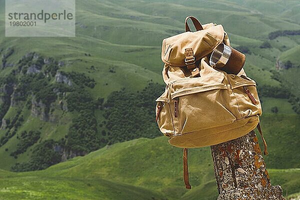 Hipster gelben Vintage Rucksack mit einem Becher auf sie mit einem Becher Nahaufnahme Vorderansicht befestigt. Traveler's Reisetasche im Hintergrund einer Berglandschaft