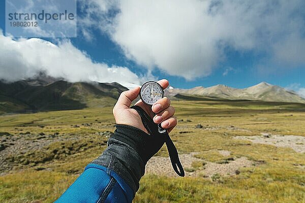 Blickpunktaufnahme. Eine Ichperspektive eines Mannes  der einen Kompass vor dem Hintergrund einer epischen Landschaft mit Klippen  Hügeln und einem blaün Himmel mit Wolken hält