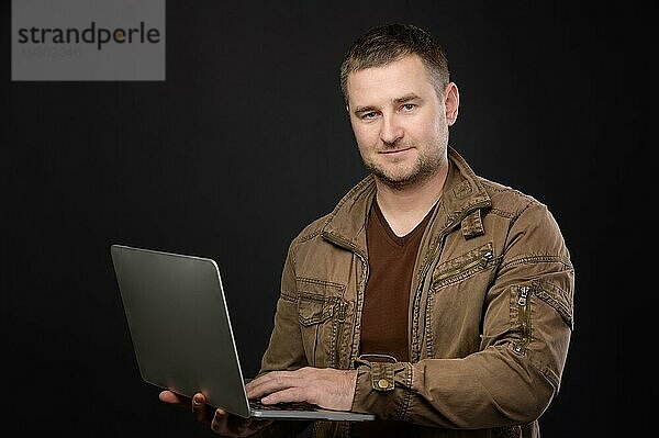 Porträt eines freundlichen  positiven  kaukasischen Mannes  der einen grauen Laptop benutzt  während er im Studio steht und in die Kamera schaut