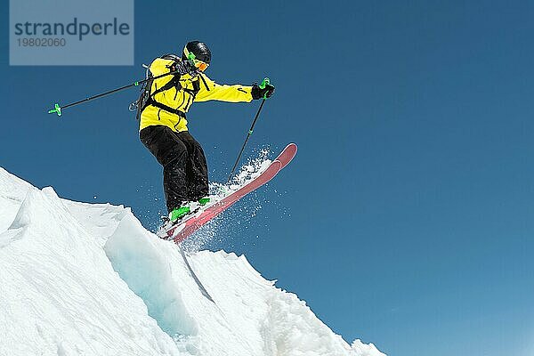 Ein Skifahrer in voller Sportausrüstung springt von der Spitze des Gletschers in den Abgrund vor dem Hintergrund des blaün Himmels und der schneebedeckten Berge des Kaukasus. Elbrus Gebiet. Russland