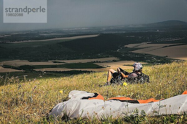 Ein professioneller Gleitschirmflieger in voller Ausrüstung und mit Helm liegt auf dem Gras hoch in den Bergen und schaut in die Wolken